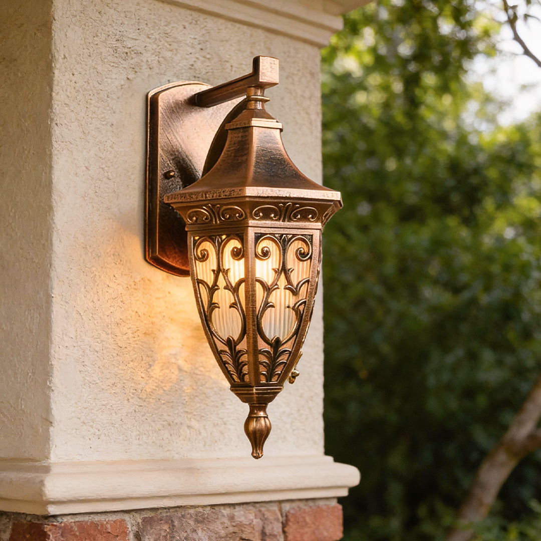 Antique art deco wall sconces with intricate bronze detailing, mounted on a beige exterior wall, emitting warm light.