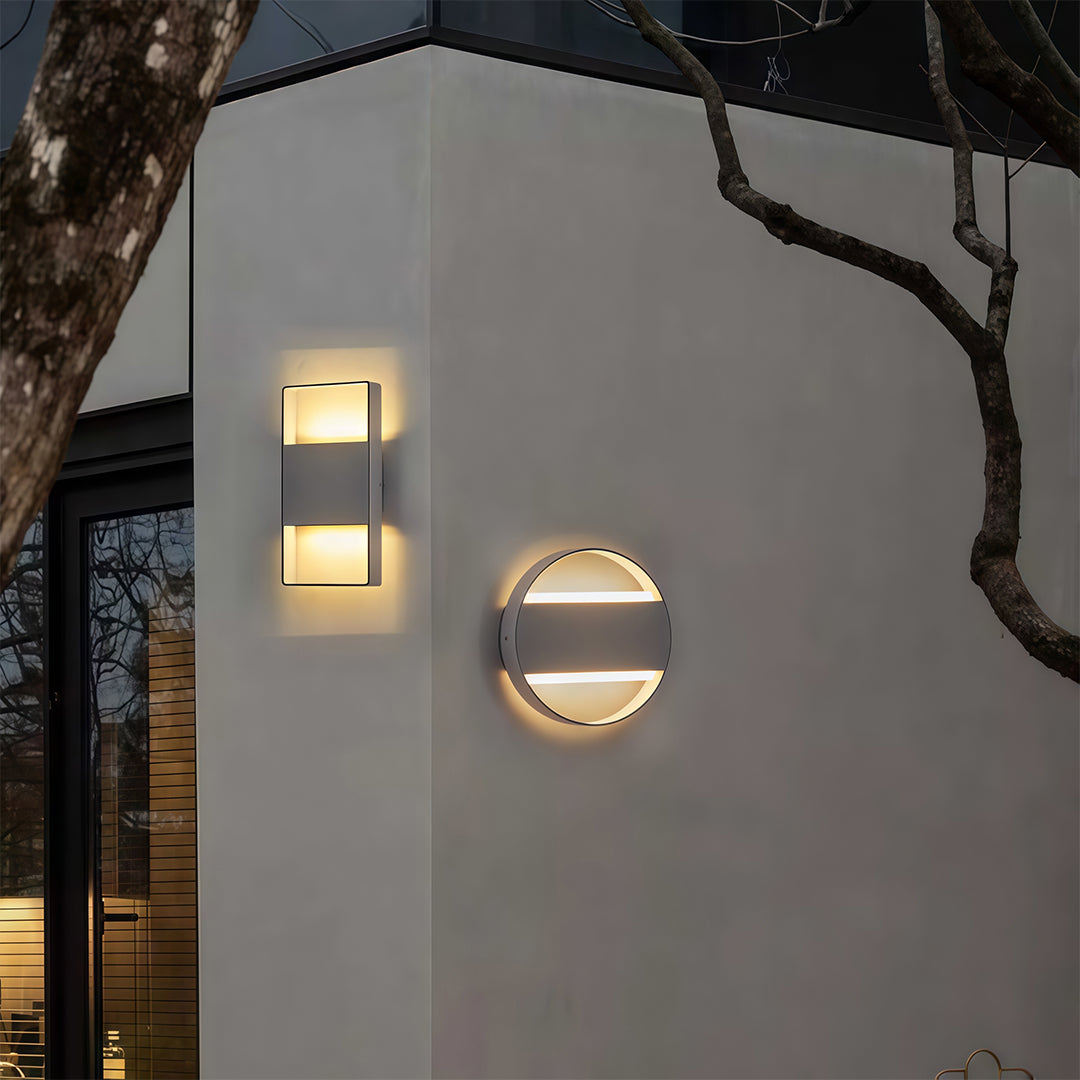 Designer mixed wall mount exterior light with glass shade chandelier styling in round and rectangular combinations on gray wall.