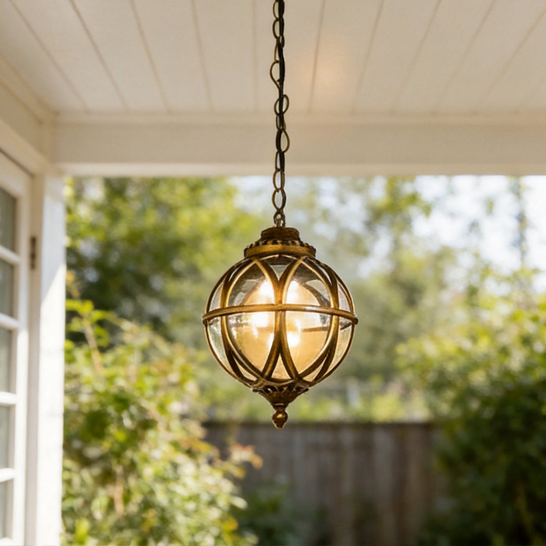 Detailed close up of a bronze metal orb pendant light hanging outdoors against blurred trees.