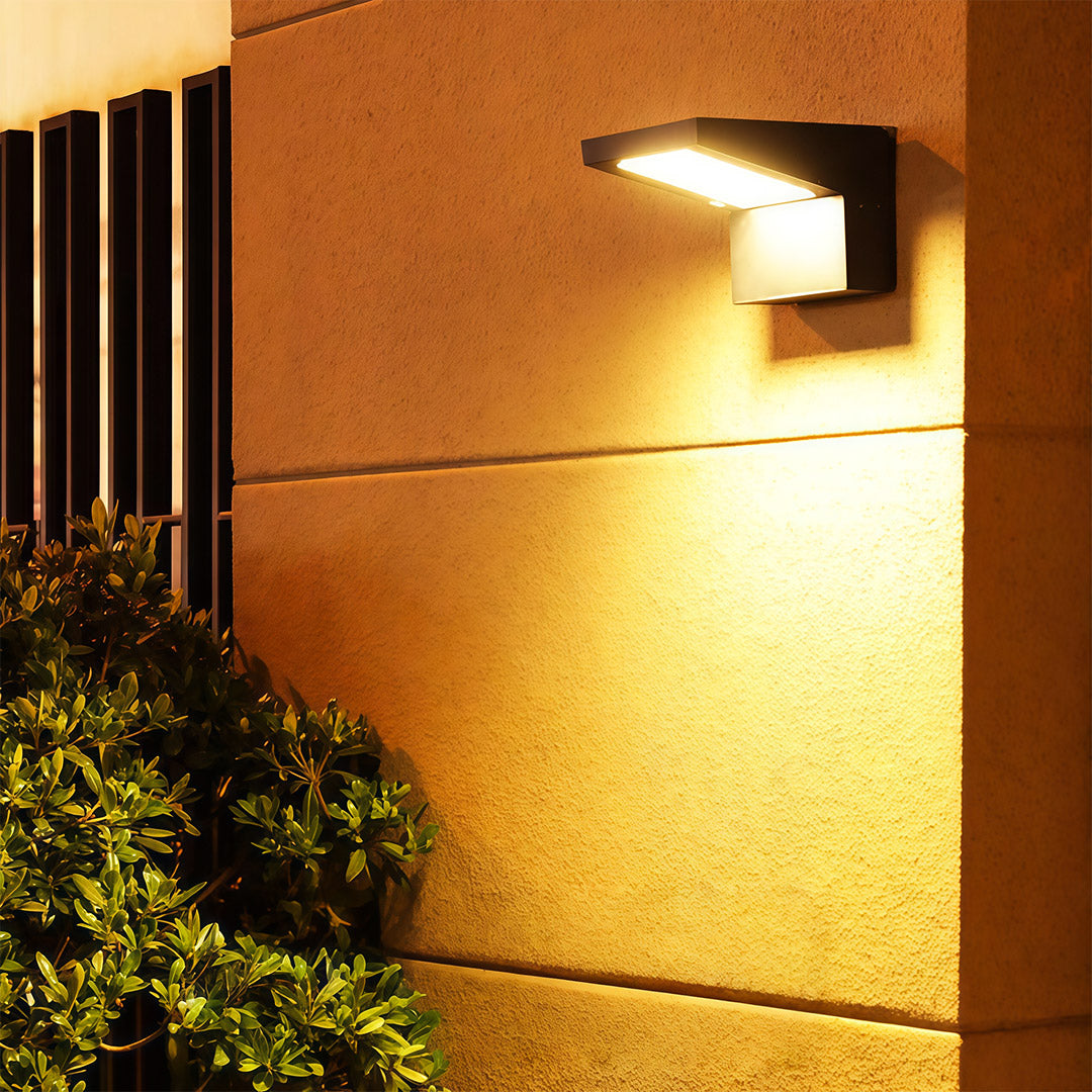 Elegant black wall mount outdoor light fixtures providing warm accent lighting above a bush on a textured wall.
