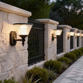 Funnel wall light with classic black finish mounted along a stone fence, illuminating a garden pathway at dusk.