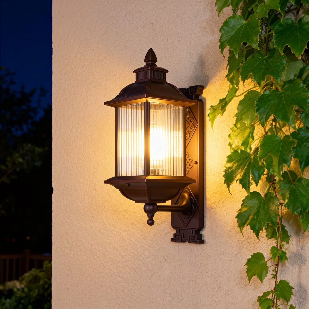 Illuminated retro outdoor wall light with warm glow mounted on textured stucco wall beside green foliage