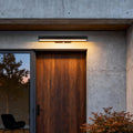 Modern LED linear wall light installed above a wooden front door, casting warm downward illumination and enhancing the minimalist exterior.