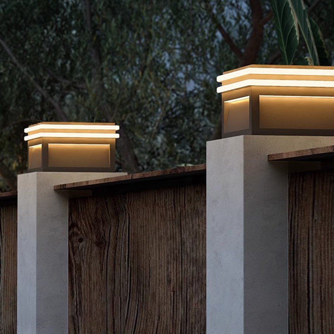 Metal pillar lights with a minimalist design, casting a soft glow on a wooden fence.