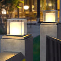 Modern gate pillar light with a sleek cube design, illuminating a garden pathway at dusk.