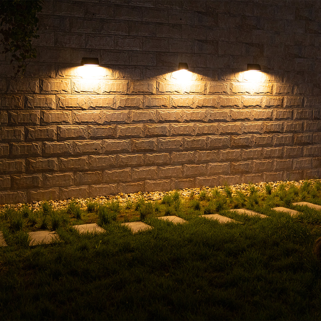 Modern outdoor wall light illuminating a brick wall and garden edge, creating a subtle nighttime landscape effect.
