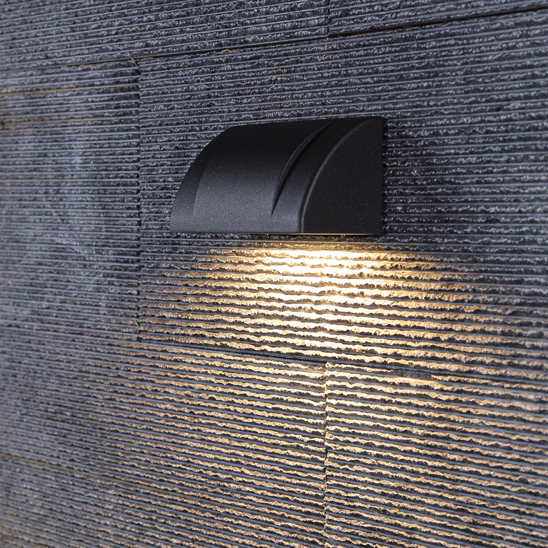 Modern outdoor wall light with a curved black housing, illuminating a stone wall with a focused downward beam.