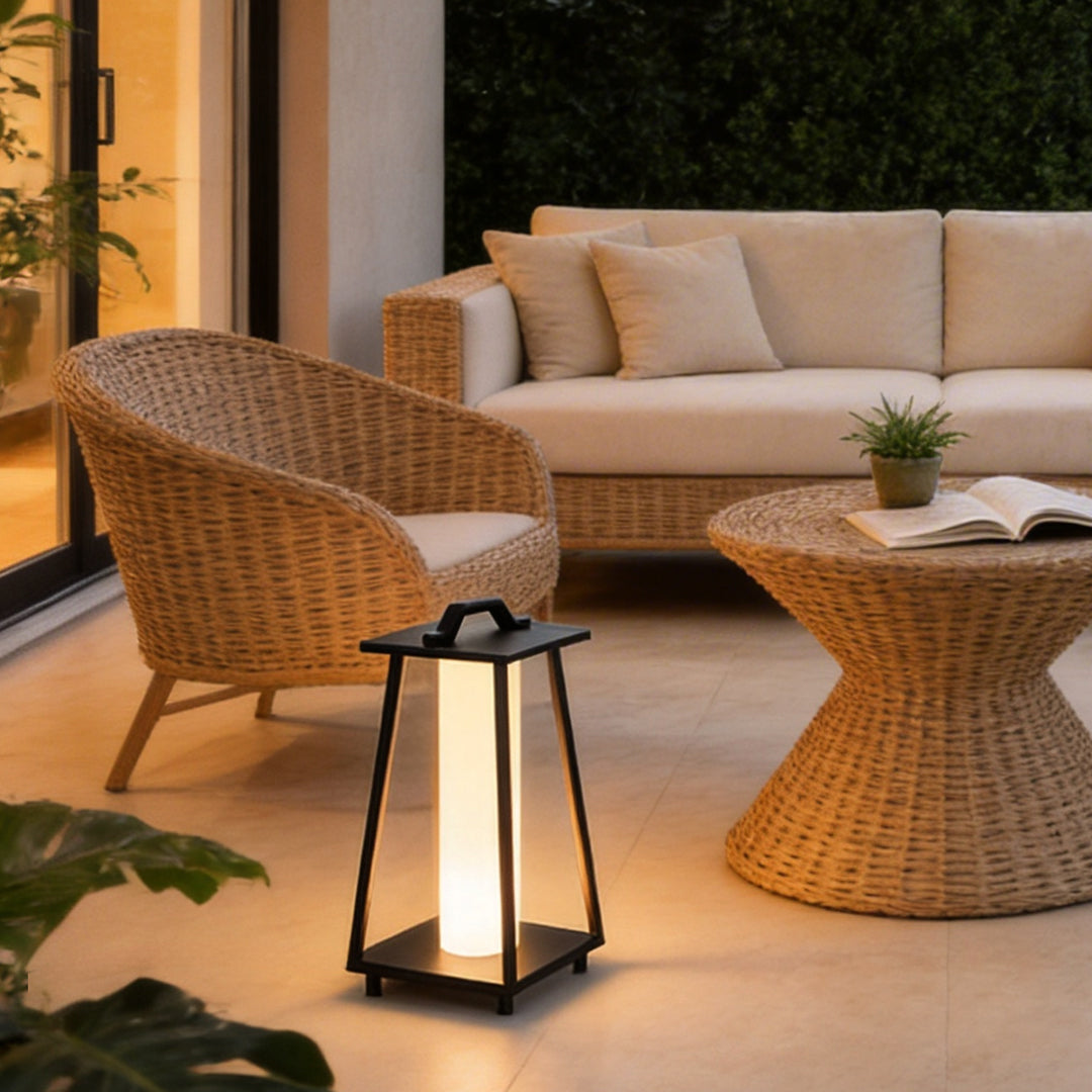 Modern portable outdoor lantern placed beside a wicker sofa on a cozy outdoor patio at night.