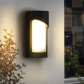 Black outdoor wall light with modern rectangular design mounted beside green plants.
