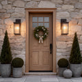 Rustic outdoor wall mounted light fixture illuminating a wooden front door with festive holiday wreath and potted plants.
