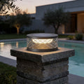 Solar cylinder pillar light with a geometric pattern, illuminating a poolside area at dusk.
