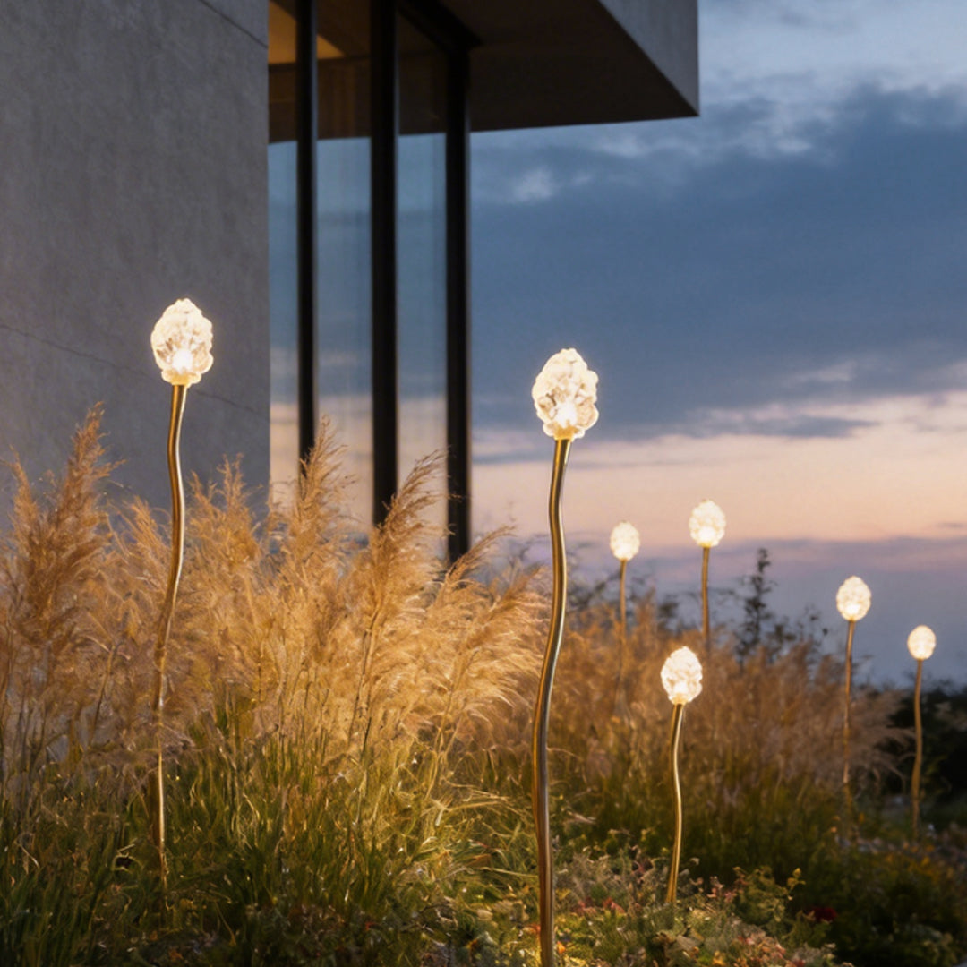 Syphasera standing lamp glowing softly among ornamental grasses beside a modern building at dusk.