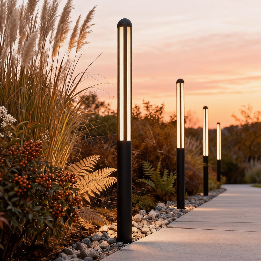 Tall garden bollard light illuminating pathway surrounded by ornamental grasses at sunset.