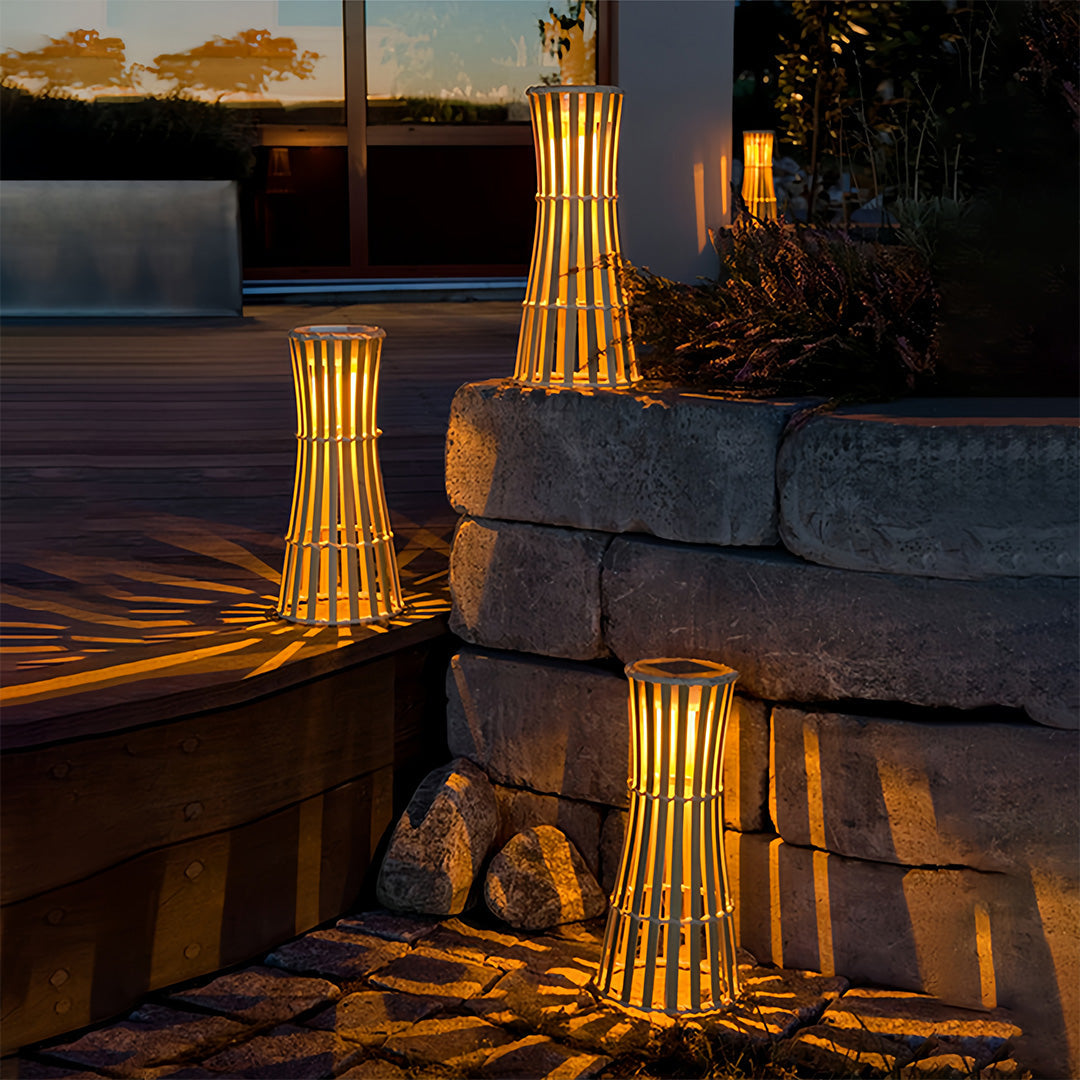 Two tall cylindrical outdoor black lanterns with vertical slat designs illuminating a wooden deck and lush green garden foliage.